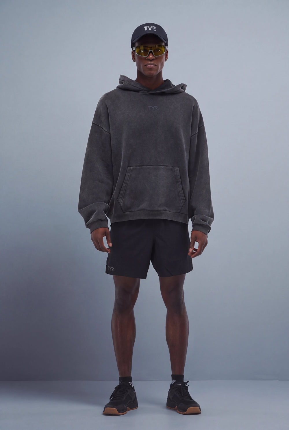 Model wearing black washed hoodie, sunglasses, black shorts, and black cxt-3 trainers.